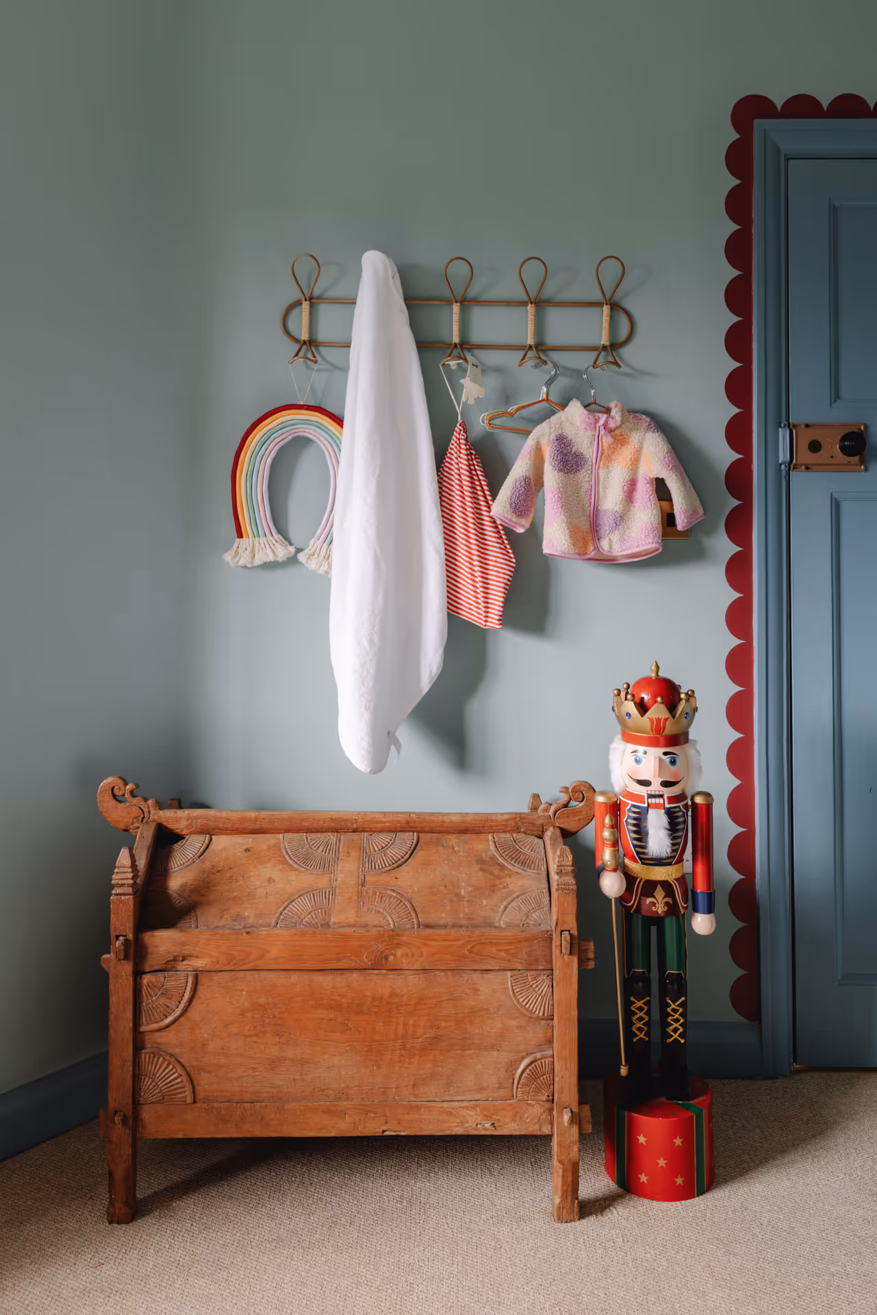 Wooden carved chest beside a large colorful nutcracker soldier in a room with light blue walls and hanging clothes and a rainbow decoration on hooks above the chest.