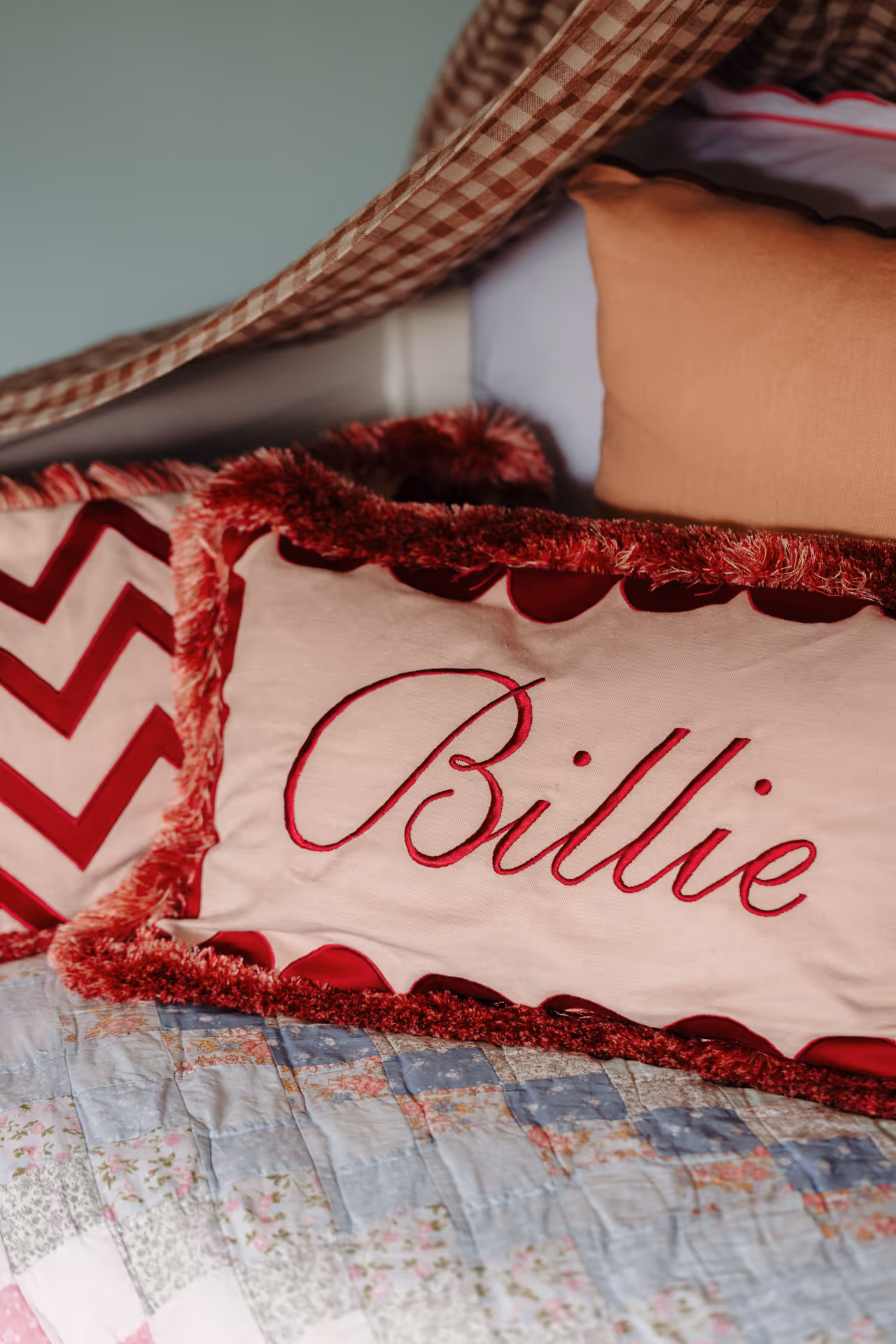 Rectangular cushion with red embroidered text 'Billie' and red fringe on a bed with patterned quilt and other cushions.