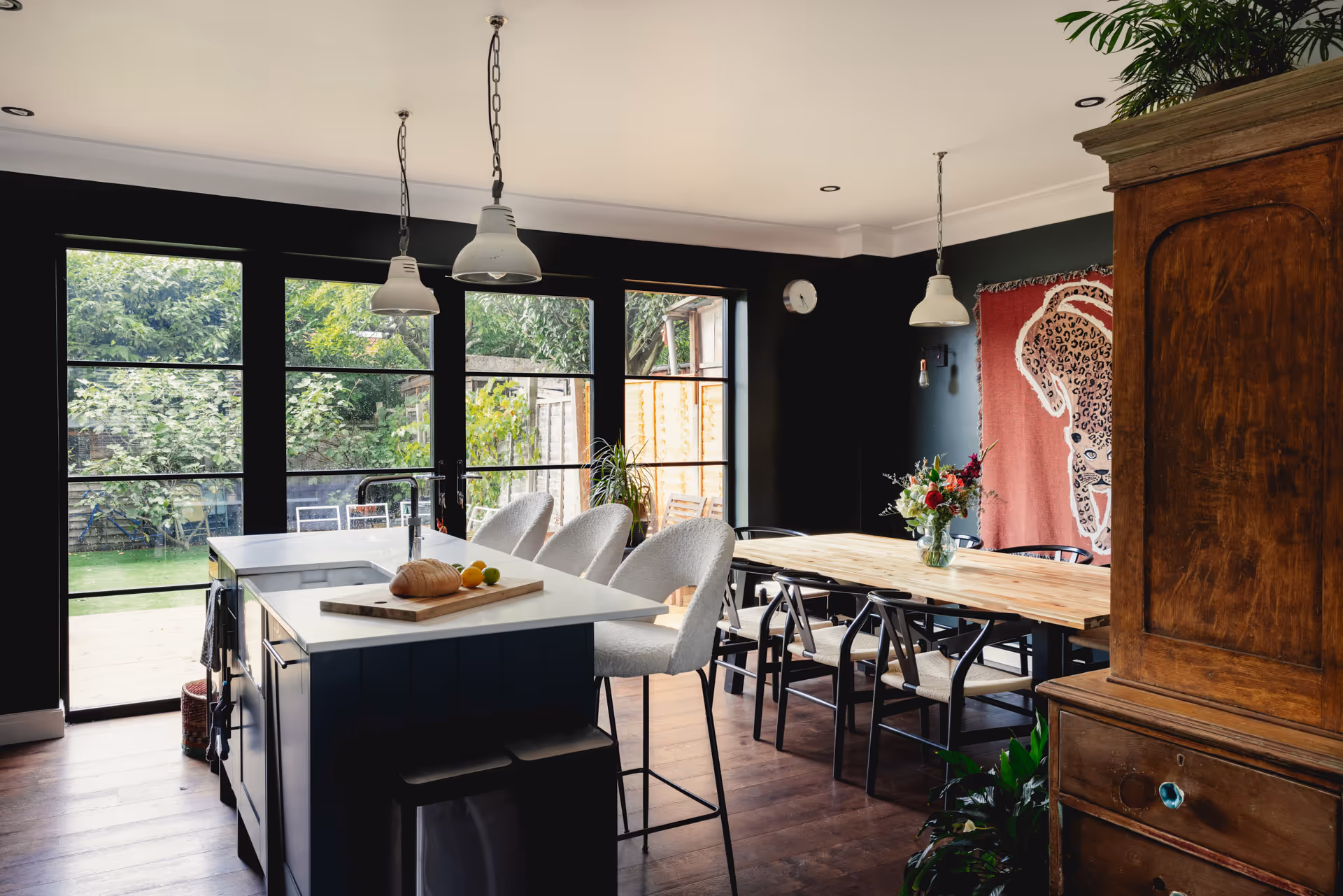 Modern kitchen diner with white countertop island, three high chairs, large wooden dining table, black chairs, and large windows overlooking a garden.