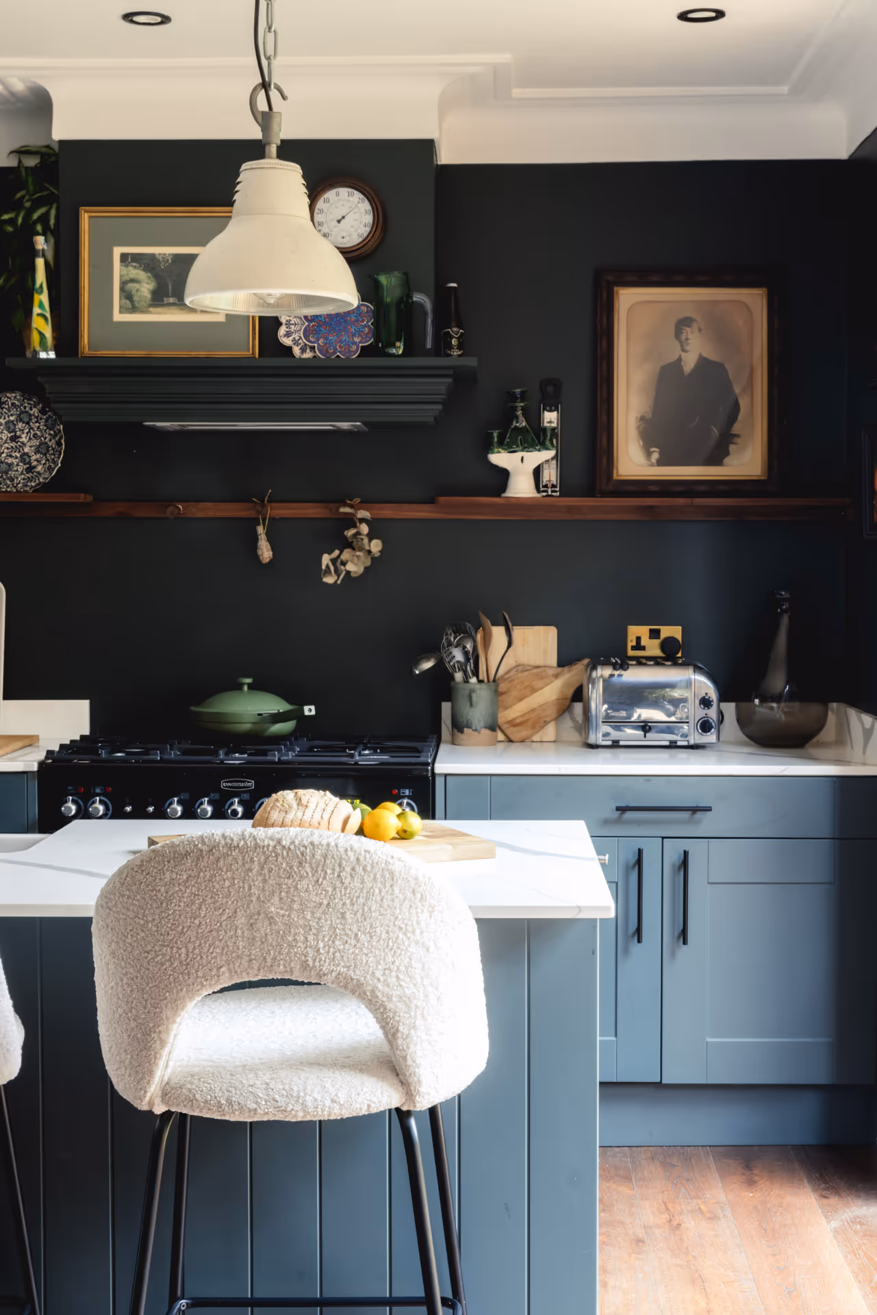 Modern kitchen with light blue cabinets, white countertop island, cream cushioned bar stool, black stove, and wall decorations including framed art and clock.