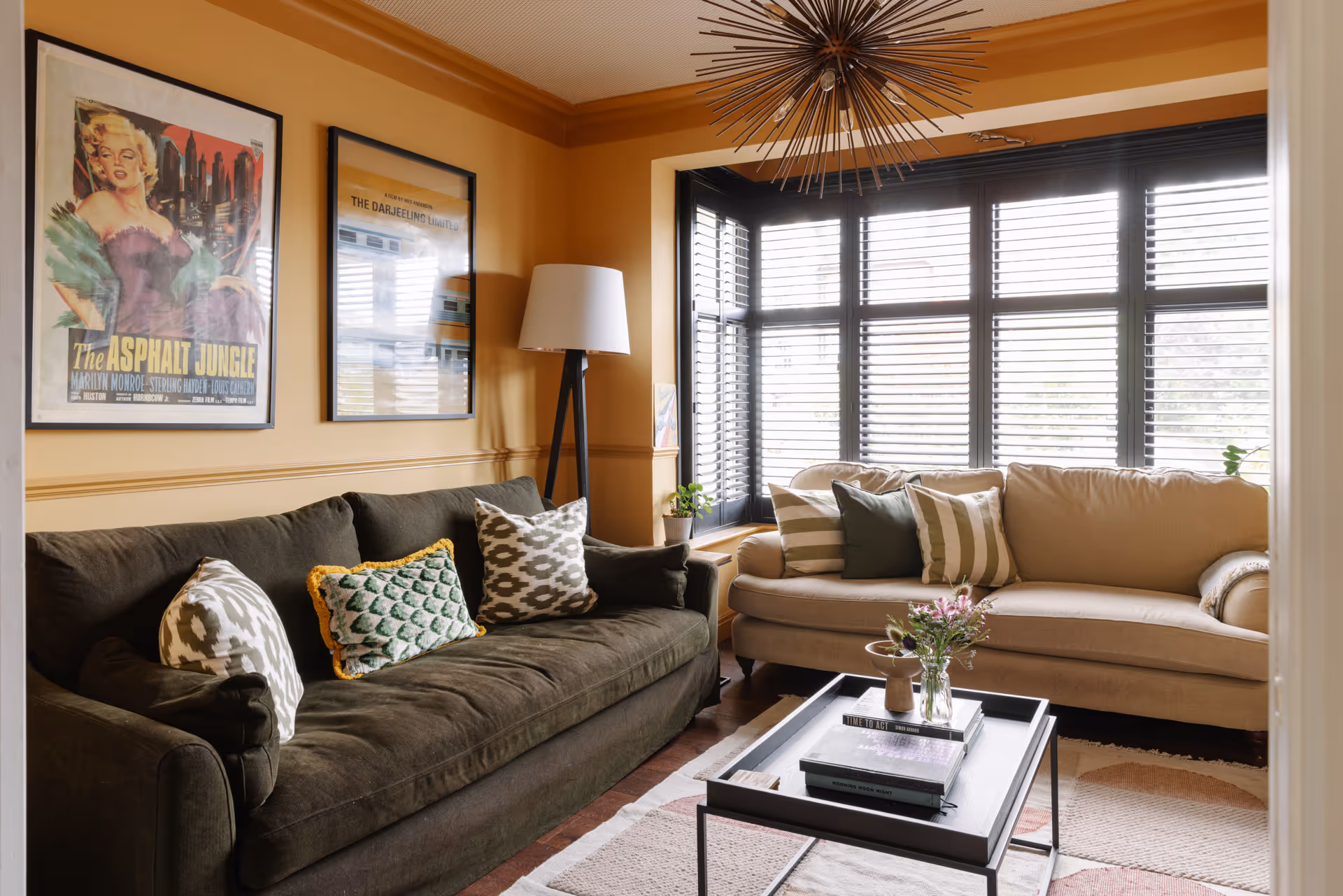 Cozy living room with a dark green sofa and beige sofa, patterned throw pillows, black coffee table with books and flowers, floor lamp, and large window with blinds.