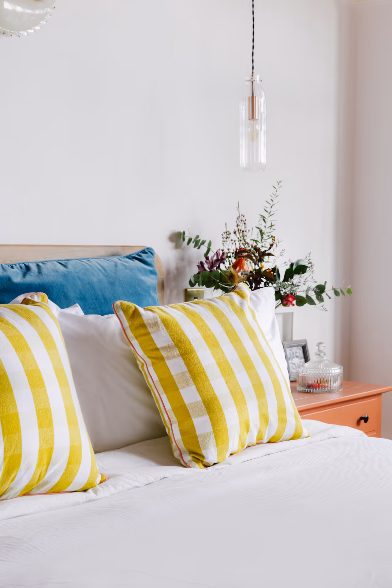 Bed with white sheets, two yellow-and-white striped pillows, and a blue pillow, next to a nightstand with flowers and a glass container.