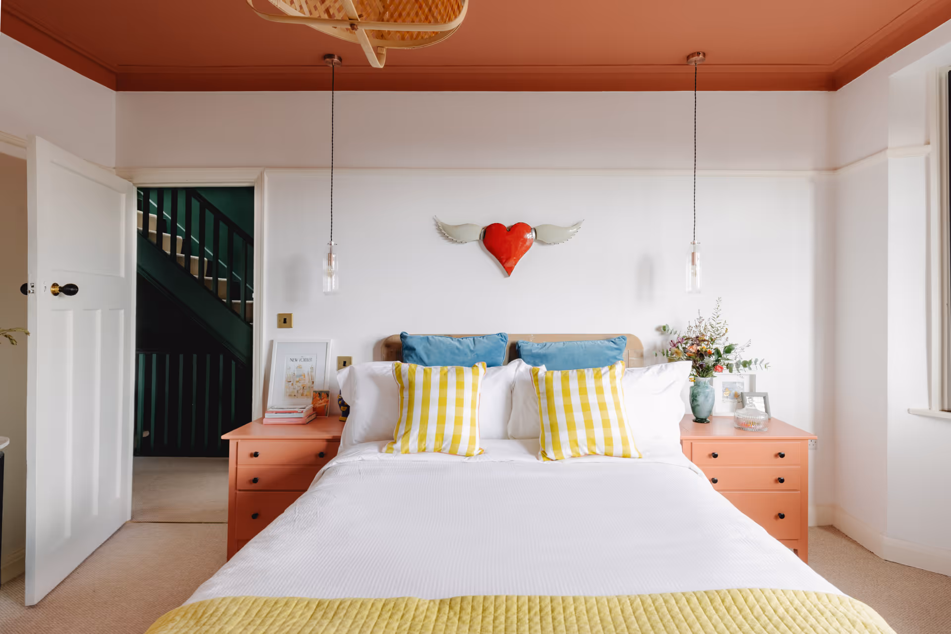 Bright bedroom with white walls, a bed with blue and yellow pillows, coral nightstands, and a red heart with wings wall decor.