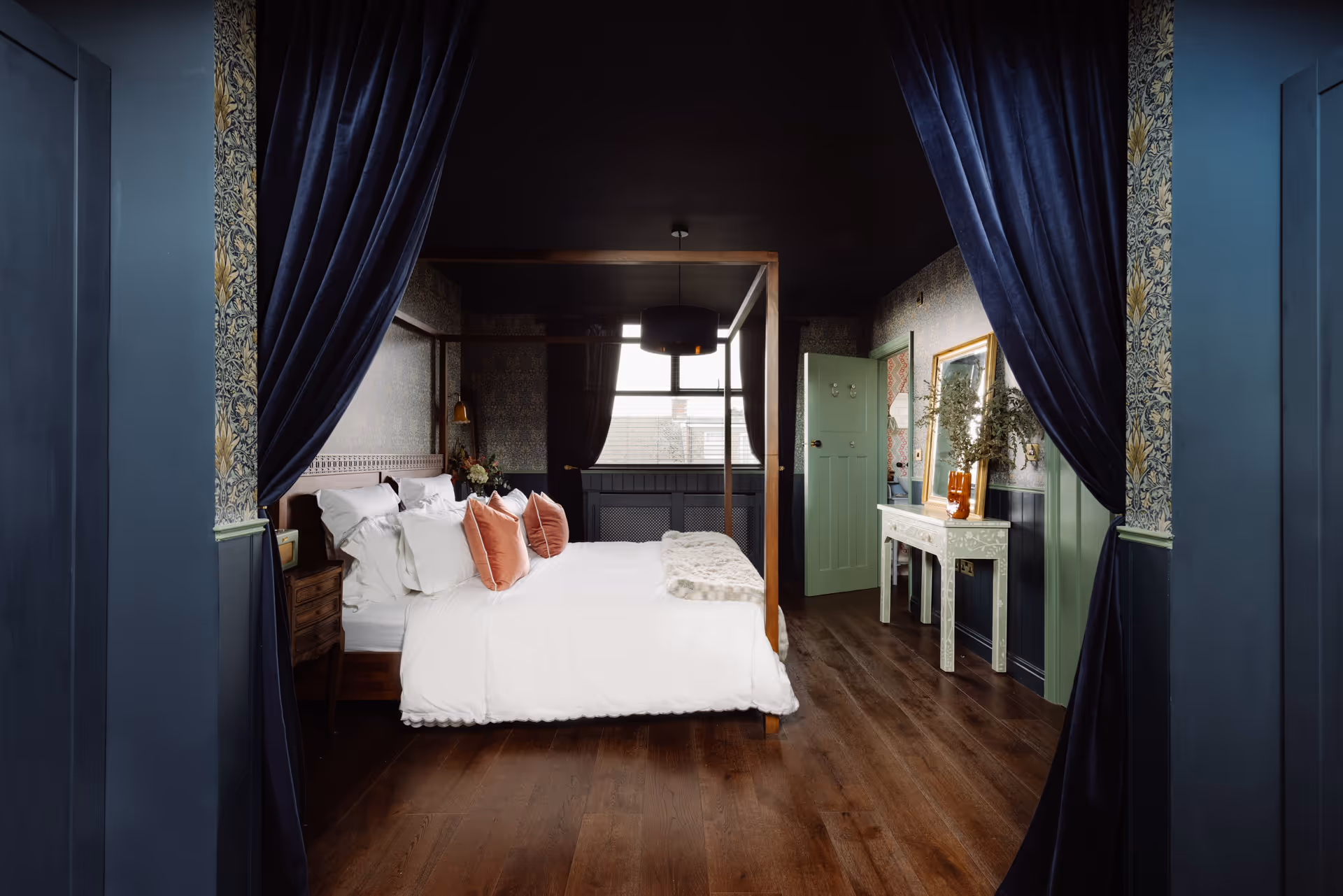 Cozy bedroom with a wooden four-poster bed dressed in white linens and pink pillows, dark walls, wooden floor, blue curtains, and a white console table with a mirror.