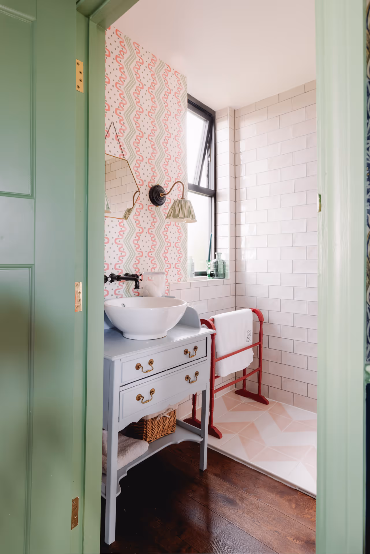 Small bathroom with light green door, patterned wallpaper, white vessel sink on a light blue vanity, and a towel rack in a tiled shower area.