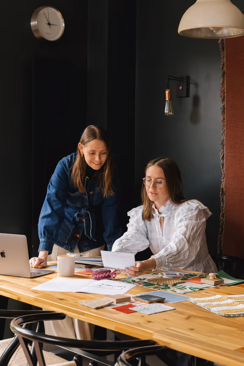 Eleanor and Jessica, the founders of House of Norica, collaborating at a wooden table with fabric samples, papers, and a laptop in a cozy, dark-walled room.
