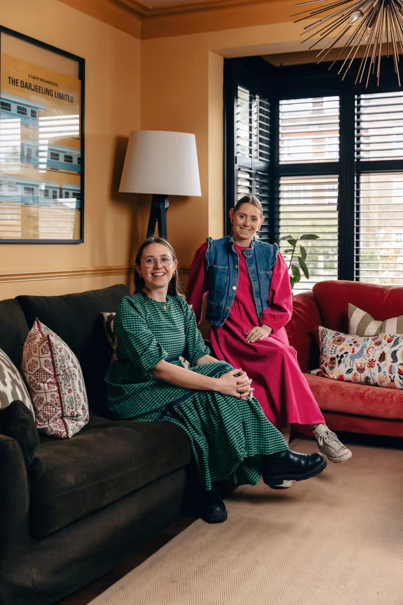 The founders of House of Norica, Eleanor and Jessica, sitting on sofas in a living room with framed art, a floor lamp, and window shutters in the background.