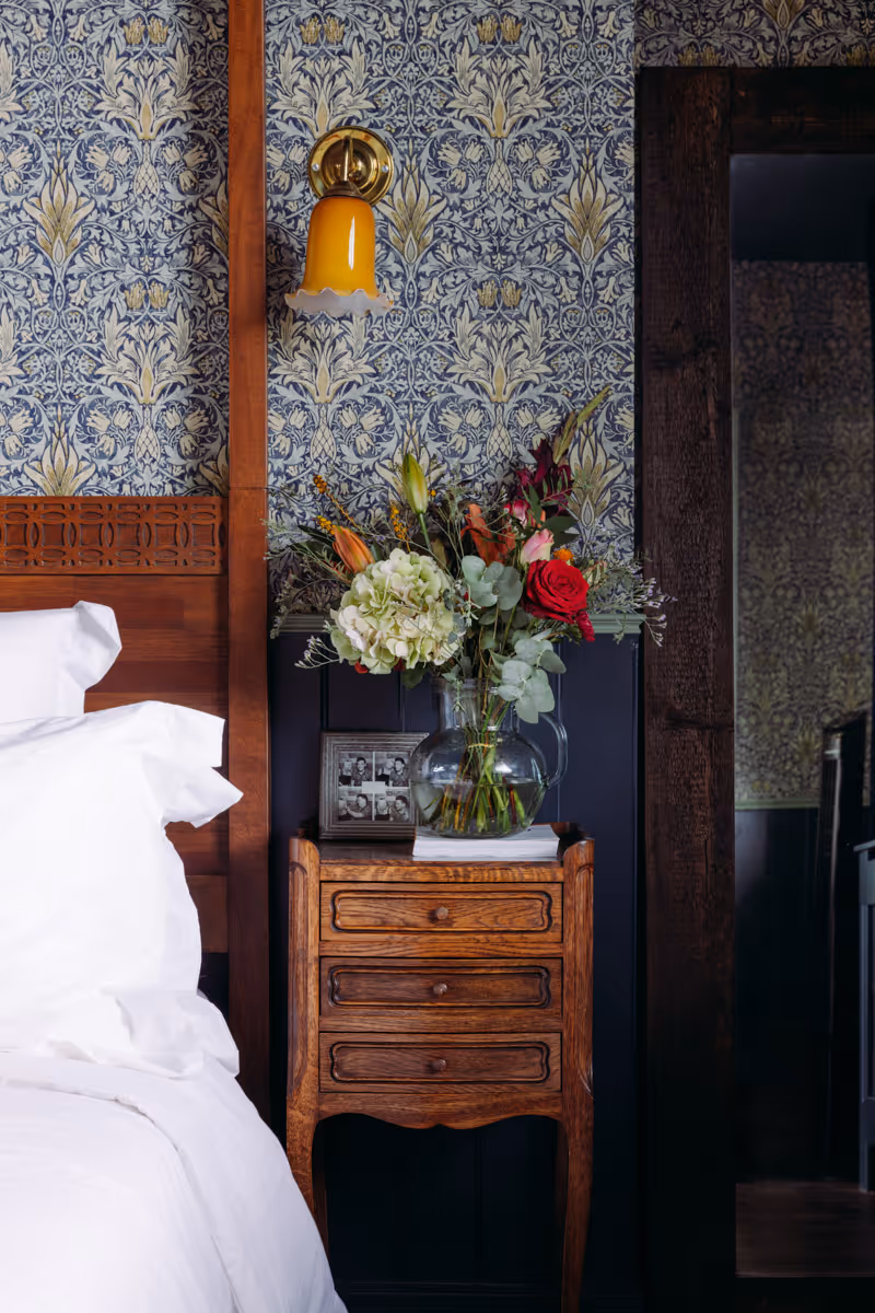 Vintage wooden bedside table with three drawers, a clear glass vase of mixed flowers, a framed black and white photo, and a yellow wall sconce above against ornate floral wallpaper.