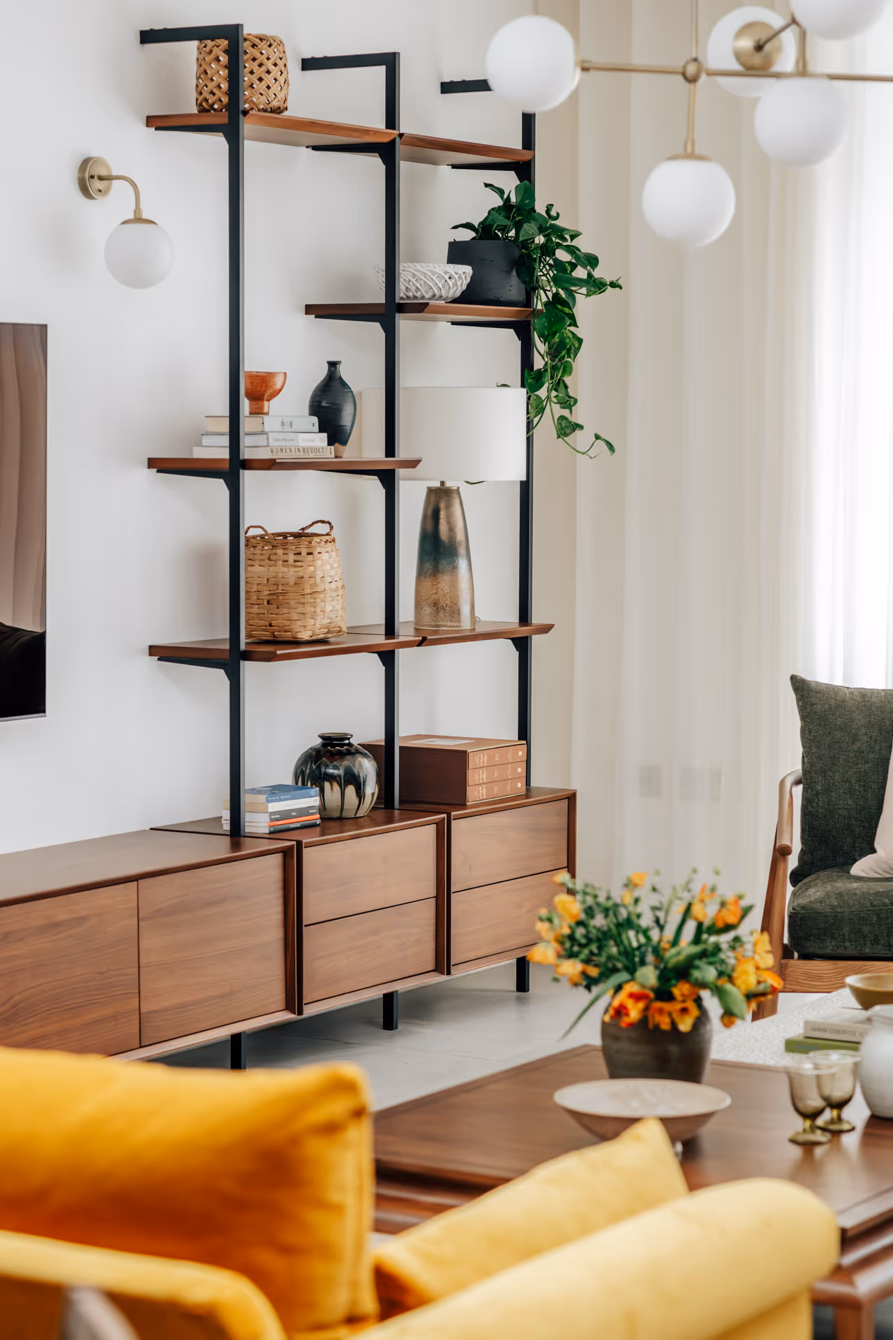 Wooden shelving unit with decorative items including woven baskets, vases, books, and a potted trailing plant in a bright living room.