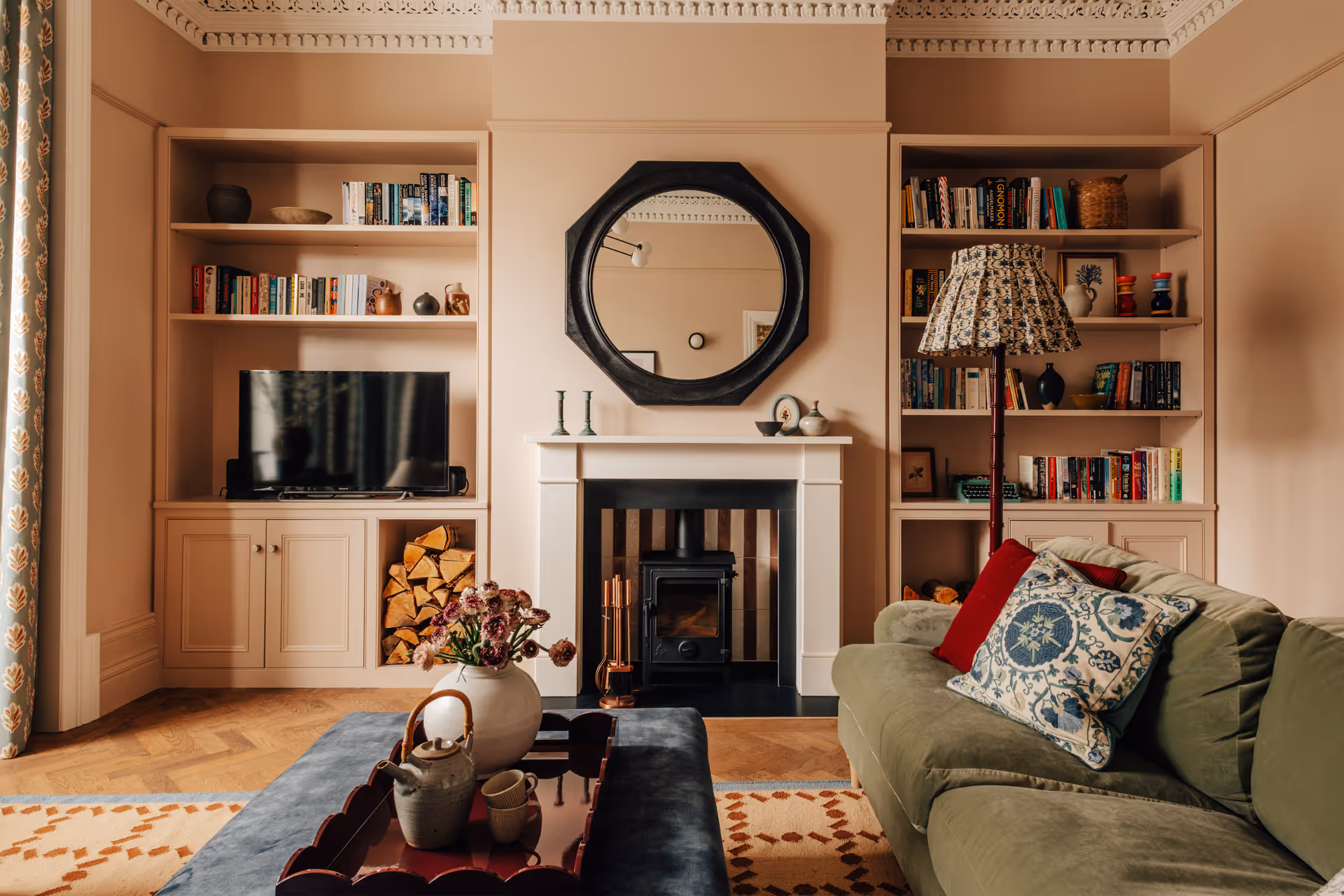 Cozy living room with beige sectional sofa, decorative pillows, round black coffee table with dried flowers and candles, green cabinet, and light wood flooring.