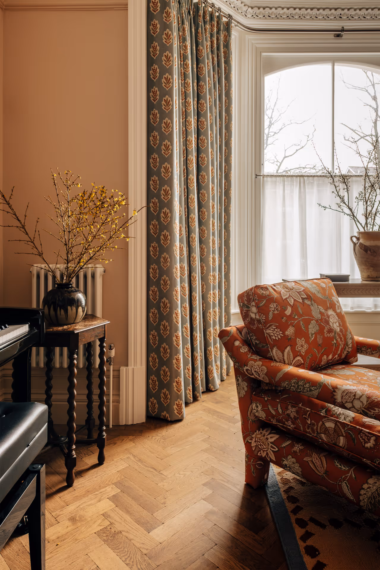 Living room corner with patterned orange armchair, wooden side table holding a vase of yellow flowers, floral curtains, and arched window with white sheer curtains.