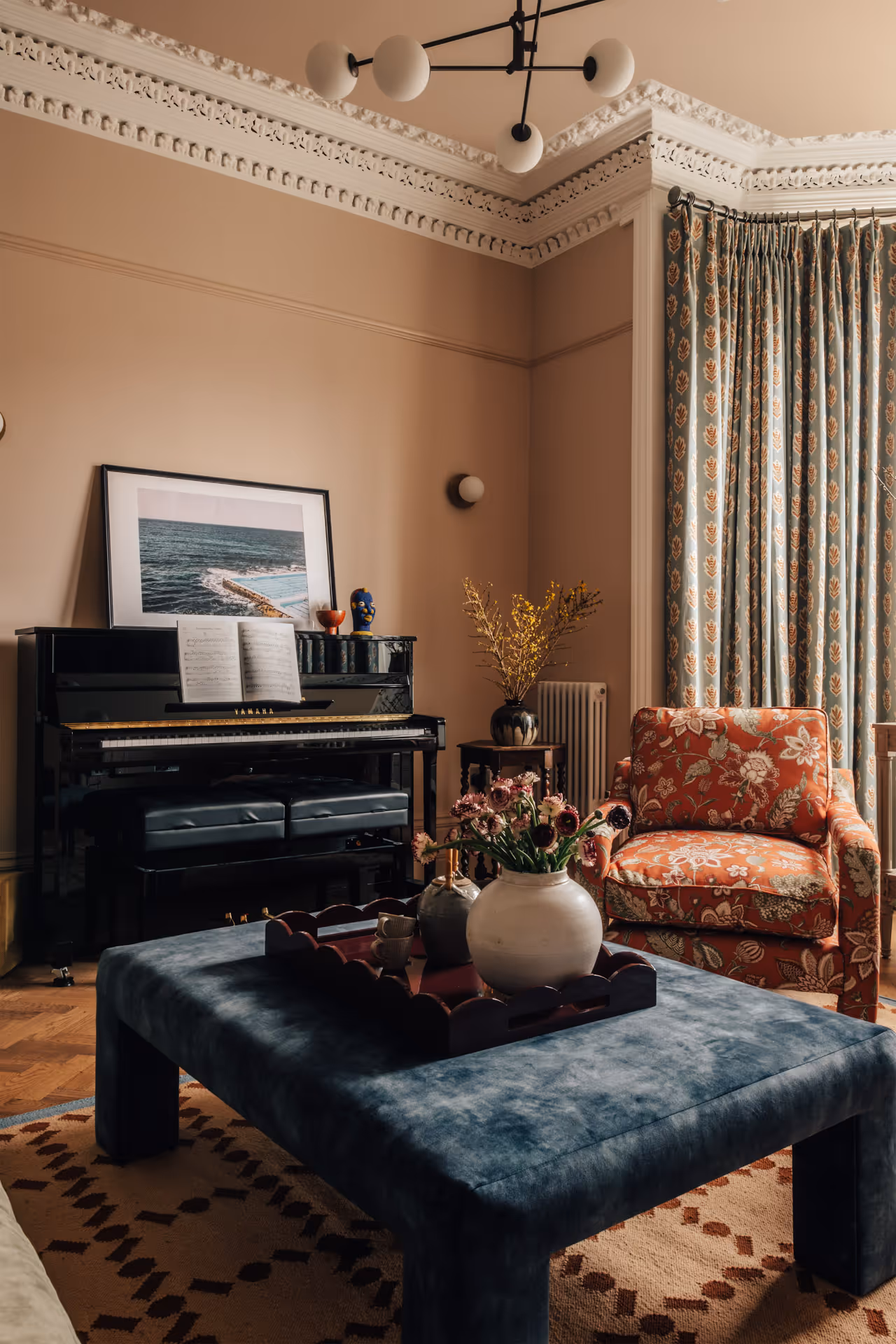 Cozy living room corner with a black upright piano holding sheet music, an orange floral armchair, a blue velvet ottoman with vases of flowers, patterned curtains, and ornate ceiling molding.