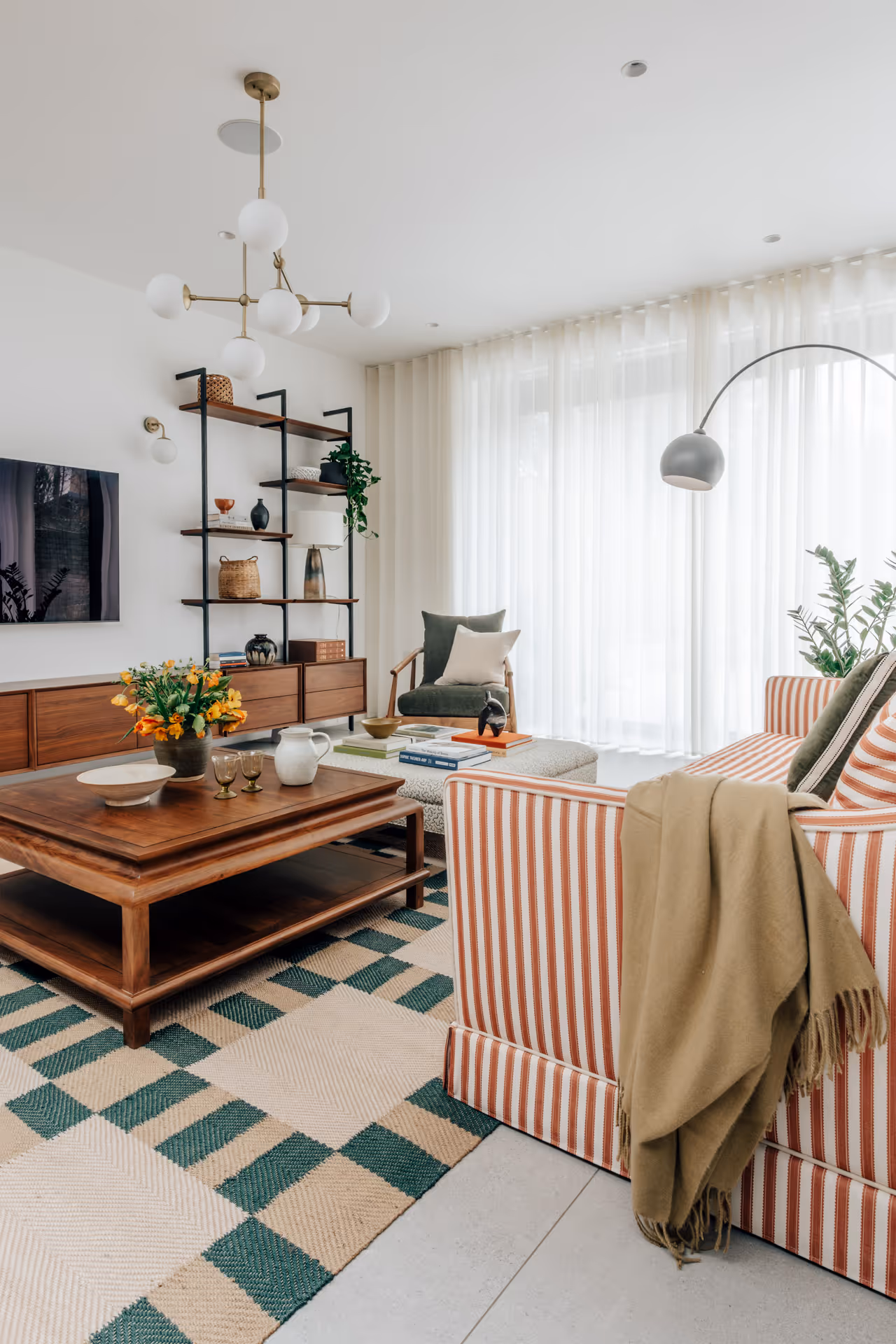 Bright living room with striped sofa, wooden coffee table, green cushioned chair, patterned rug, and large windows with sheer curtains.
