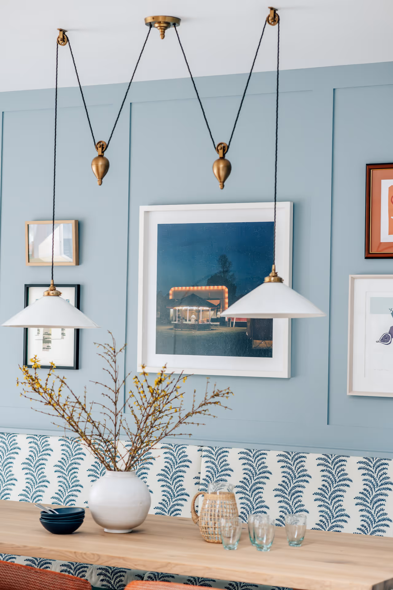 Dining area with light wooden table, white vase with branches, glassware, patterned bench, and two hanging pendant lights against a blue paneled wall with framed artwork.