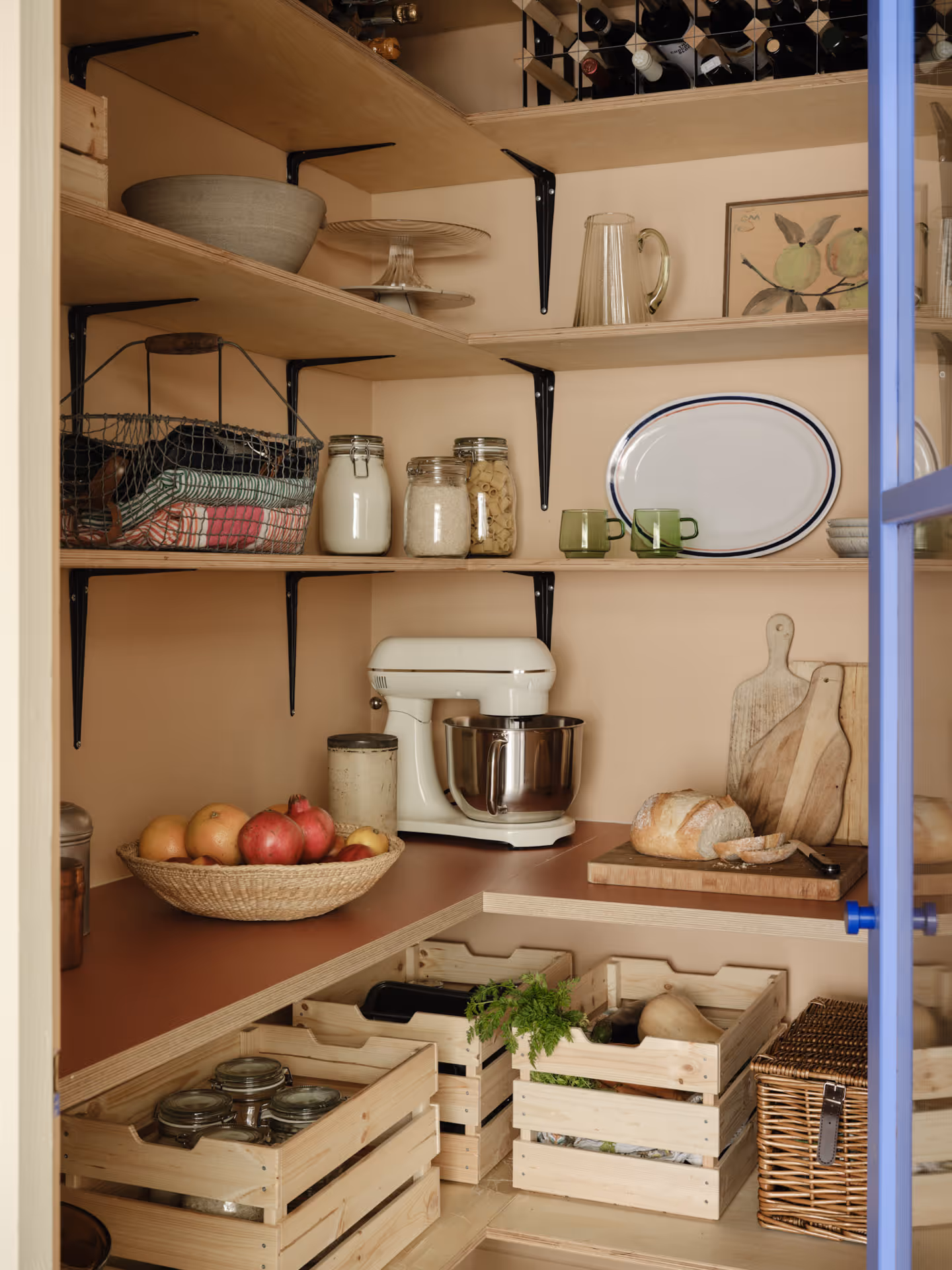 Organized pantry shelves with jars, mixing bowl, mixer, fruit basket, cutting boards, bread on a board, wooden crates with vegetables, and wine bottles on top shelf.