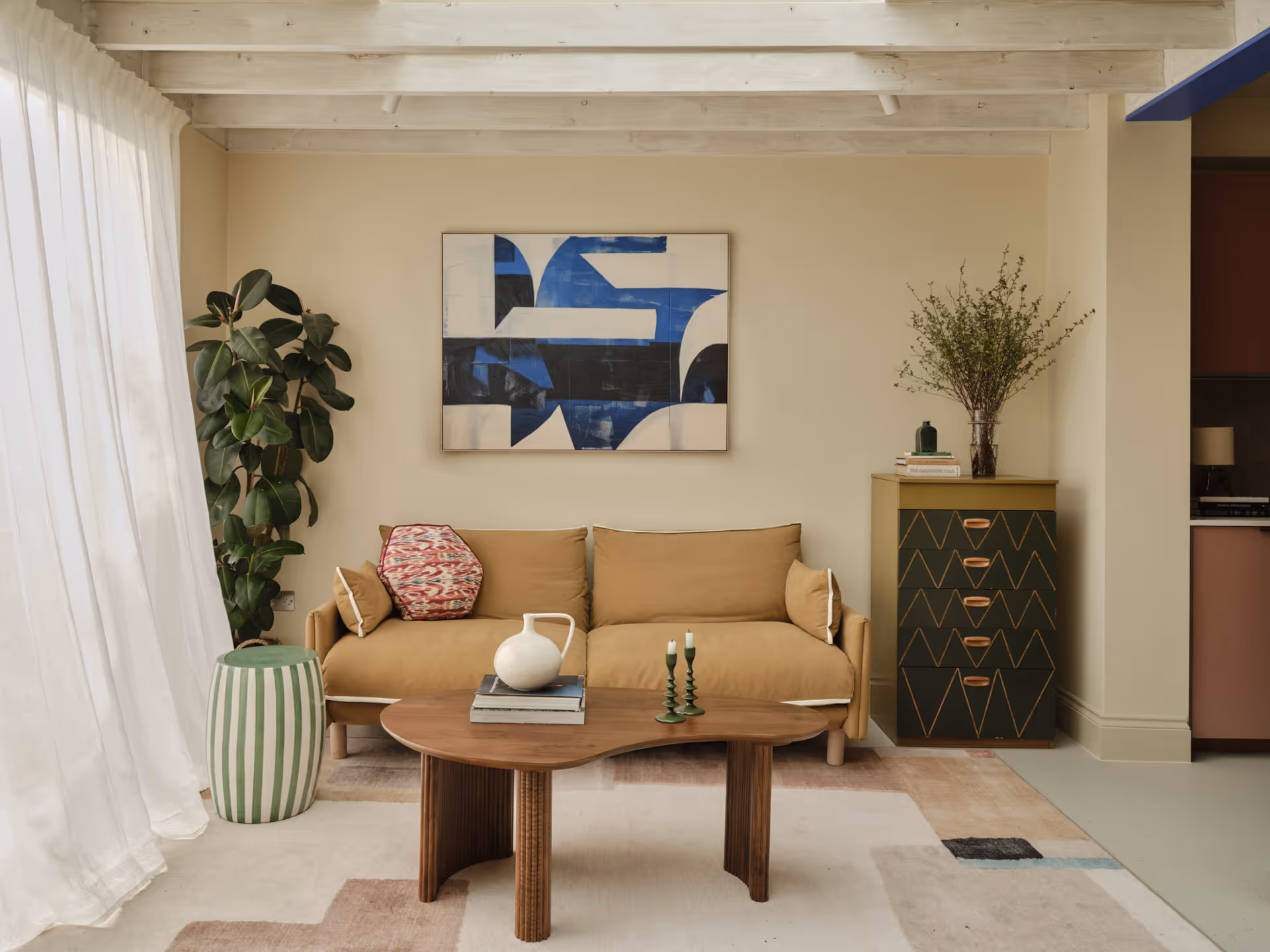 Living room with beige sofa, abstract blue and white painting, wooden coffee table with vase and candles, green-striped stool, indoor plant, and patterned cabinet with vase of branches.