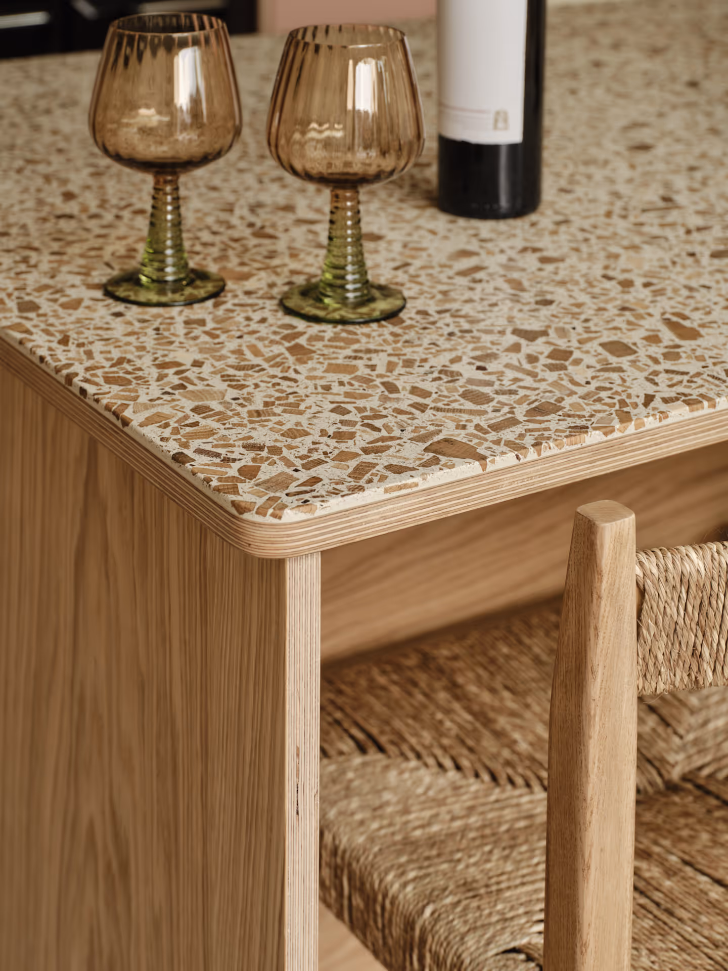 Close-up of a terrazzo countertop with two green-stemmed amber wine glasses and a black-and-white wine bottle, next to a wooden chair with woven seat.