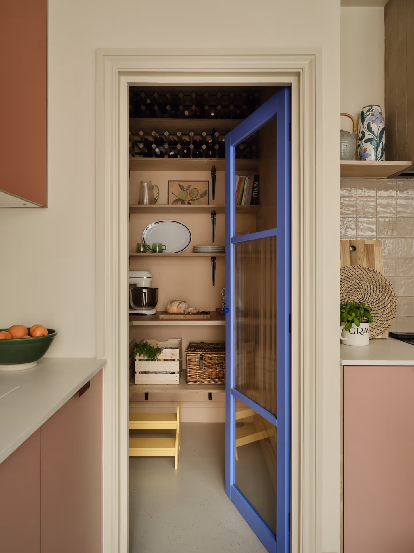 Open pantry door with blue frame showing shelves stocked with wine bottles, kitchenware, bread, and storage baskets.