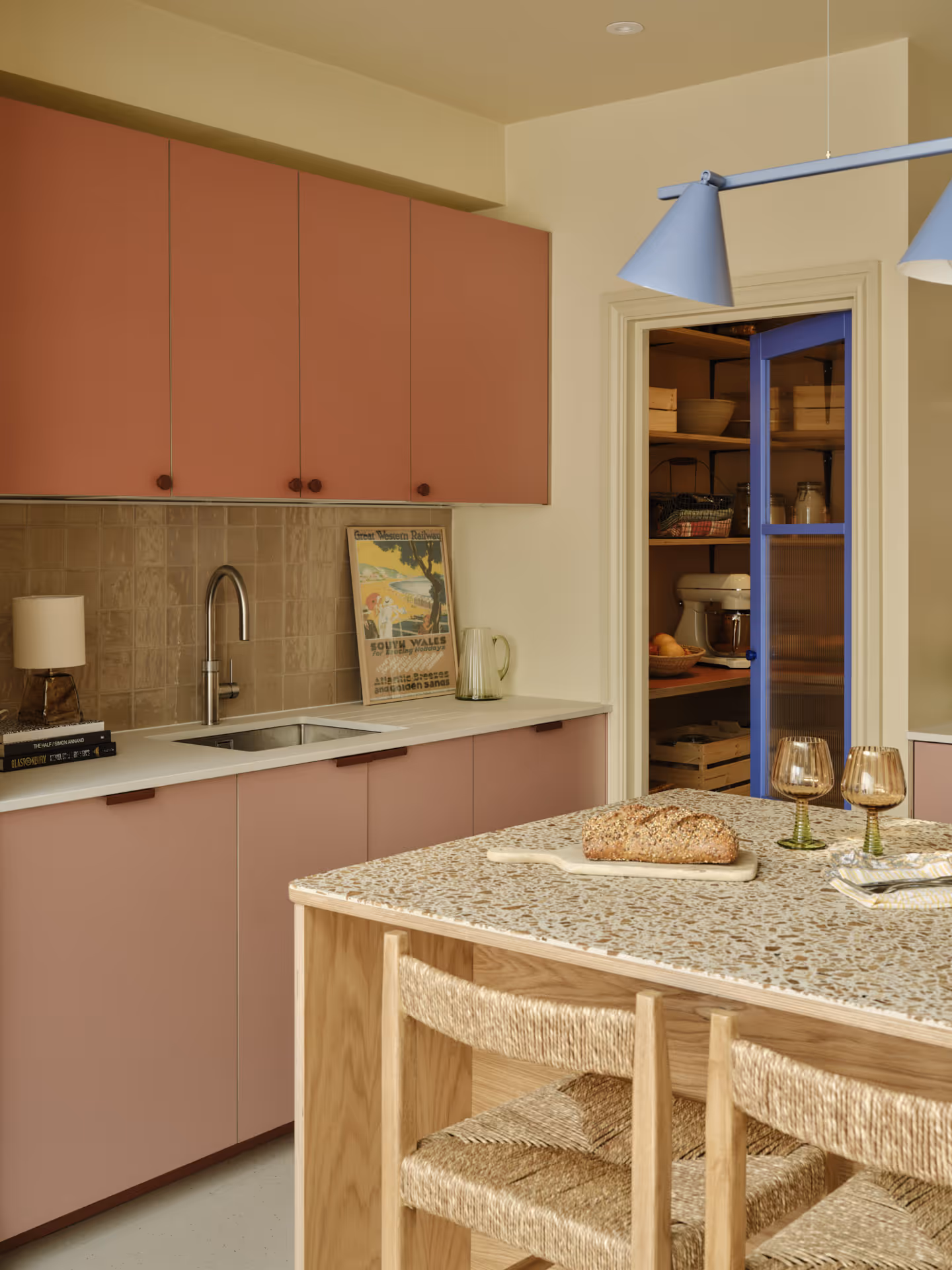 Modern kitchen with pink cabinets, terrazzo countertop island with woven chairs, loaf of bread, two wine glasses, and a pantry with a blue door.