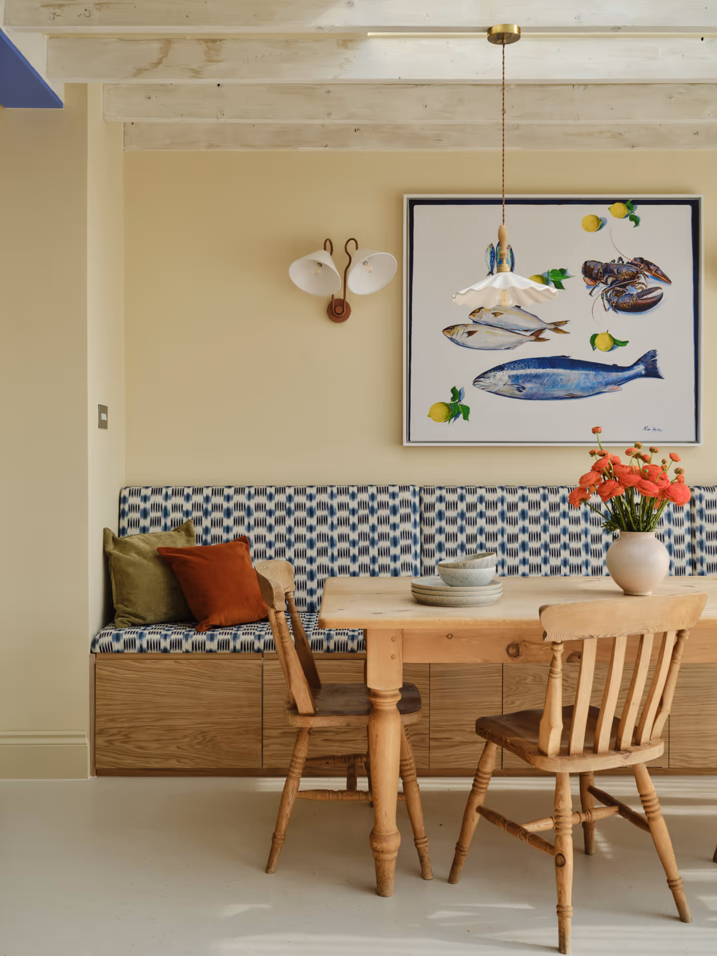 Cozy dining area with a wooden table, two wooden chairs, a patterned bench with cushions, floral centerpiece, and wall art featuring fish and lemons.