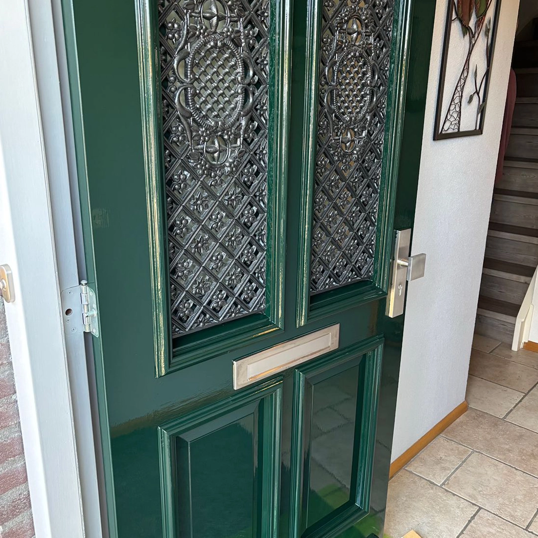Donkergroene voordeur met sierlijk bewerkte metalen raampanelen en zilverkleurig deurbeslag.