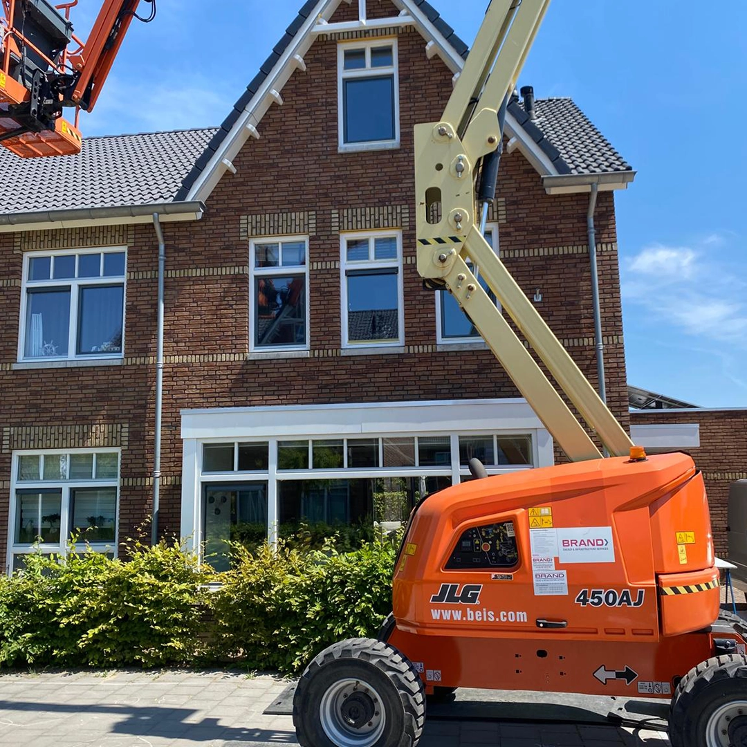 Hoogwerker voor een traditioneel huis met roodbruine gevel; schilders zijn bezig met schilderwerk op hoogte.