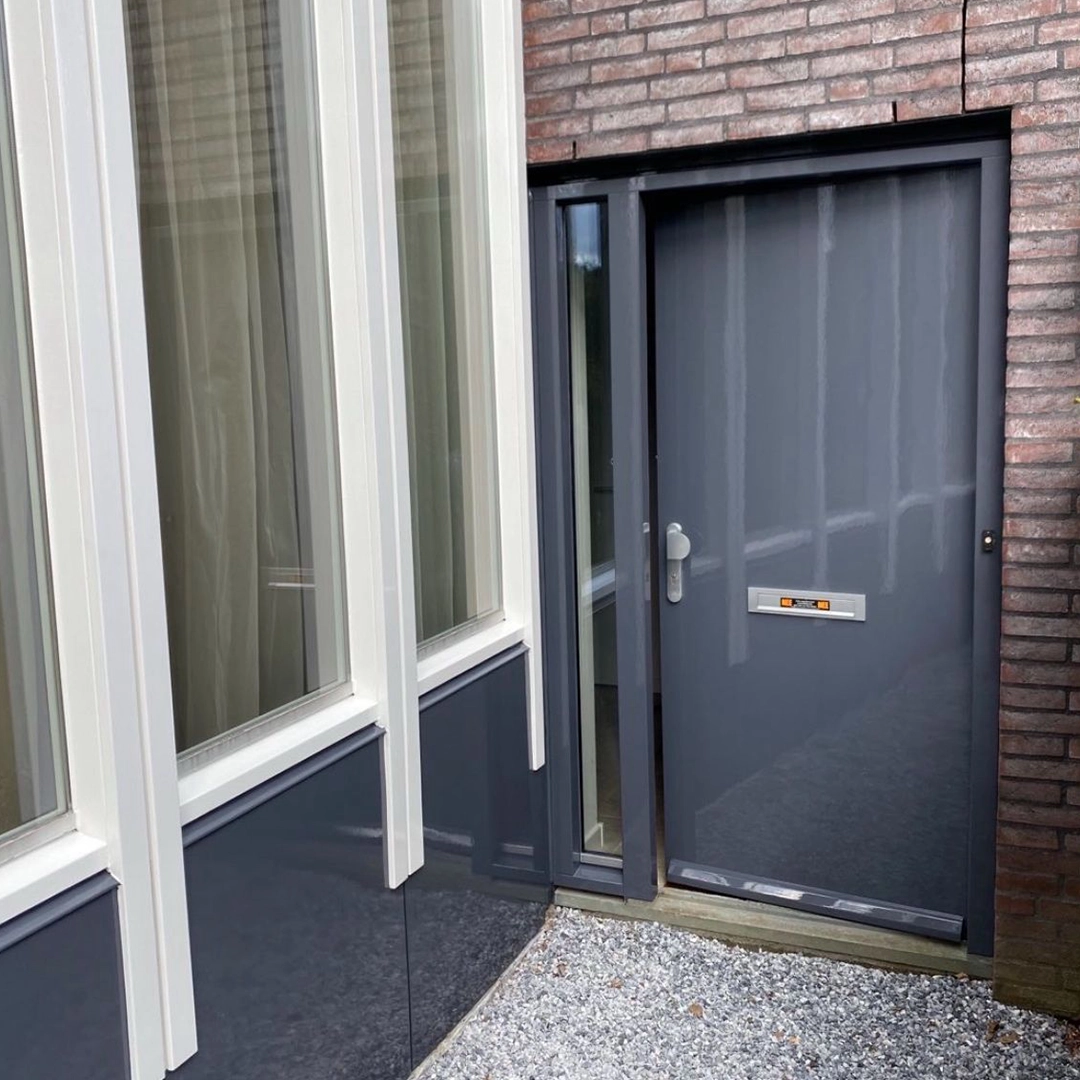 Grijze voordeur met glaspaneel naast witte kozijnen en grijze gevelbekleding, aan een bakstenen woning.