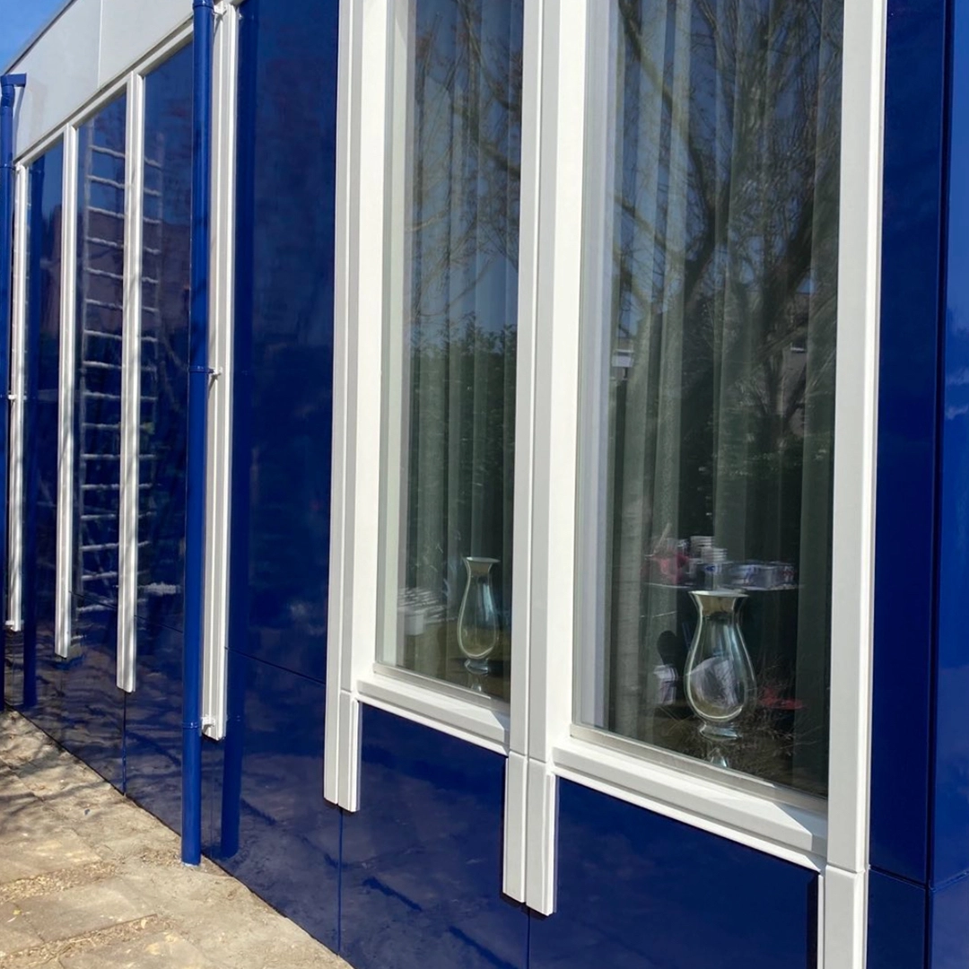 Blauw glanzend geschilderde gevel met witte kozijnen en regenpijpen, voorzien van grote ramen.