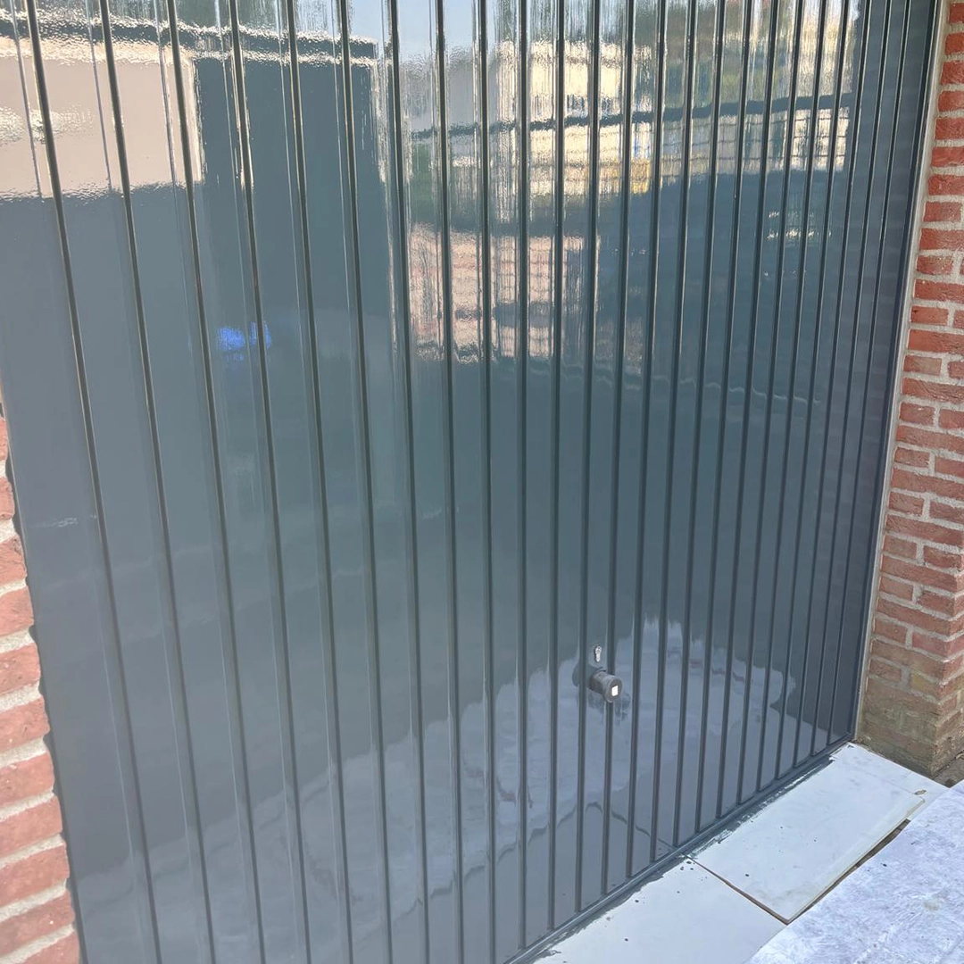 Donkergrijs geschilderde garagedeur met verticale banen, strak afgewerkt tegen een bakstenen muur.