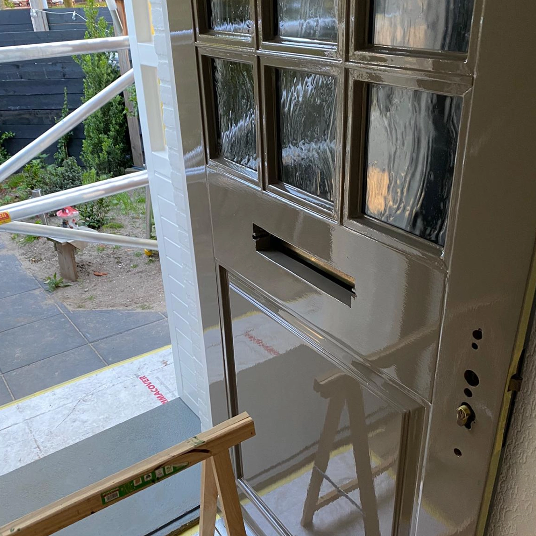 Glanzend gelakte bruingrijze voordeur met klassiek glas en witte omlijsting.