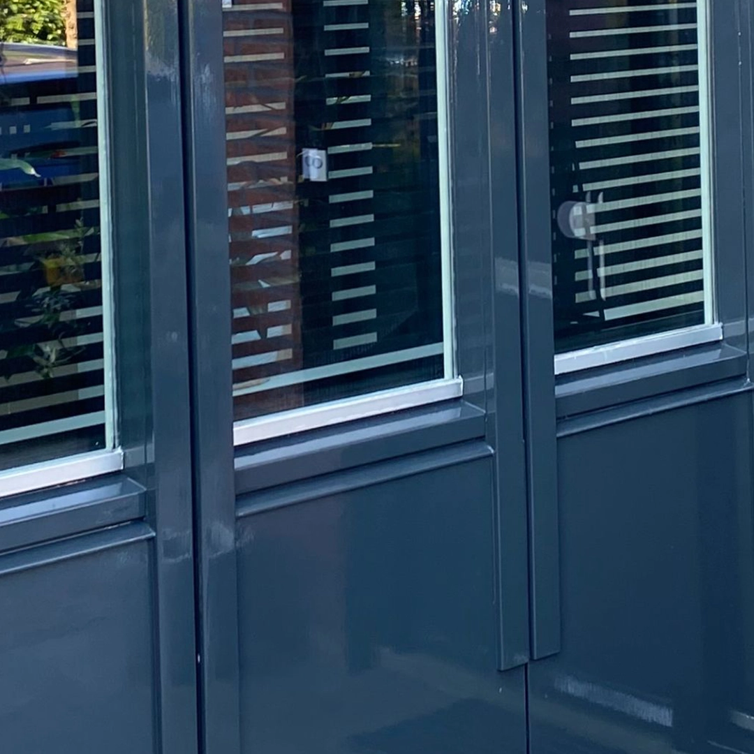 Close-up van donker geschilderde gevel met ramen voorzien van witte kaders en jaloezieën.