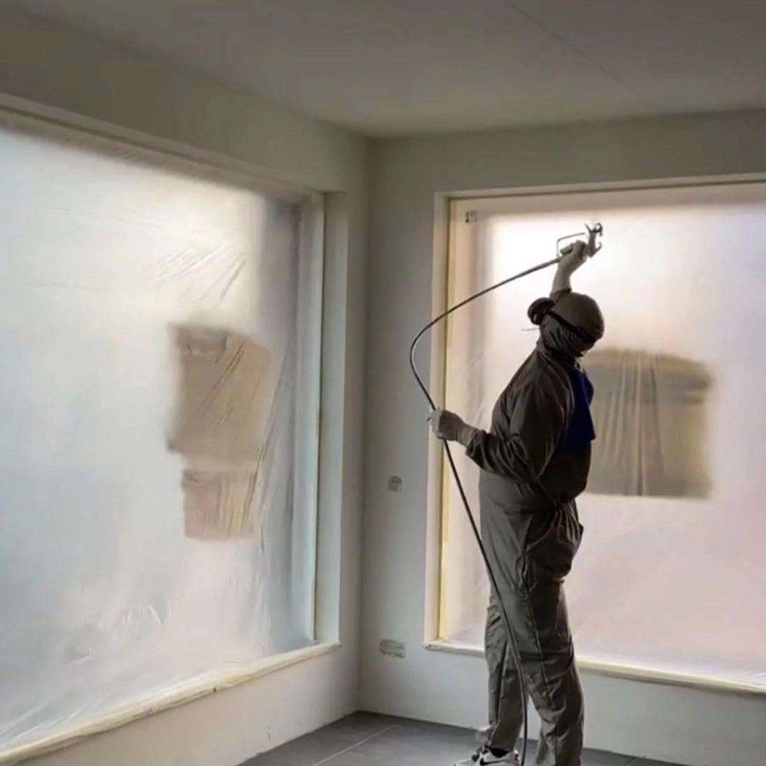 Schilder in beschermende kleding spuit verf tegen een muur in een ruimte met afgeplakte ramen en grijze vloer.