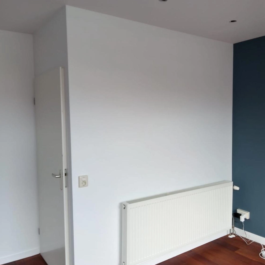 Strak wit geschilderde wand met een donkerblauwe accentmuur, witte radiator en deur, en een houten vloer voor een warme afwerking.
