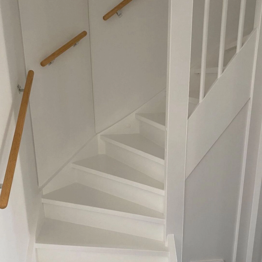 Deze trap is strak wit afgewerkt, met een moderne uitstraling en een cleane afwerking die mooi past in een licht interieur. De combinatie met houten leuningen zorgt voor warmte en contrast, waardoor het geheel vriendelijk en uitnodigend oogt. Ook hier is zichtbaar dat er netjes is gewerkt: de lijnen zijn zuiver, zonder druipers of vlekken. Een verfijnde, frisse look.