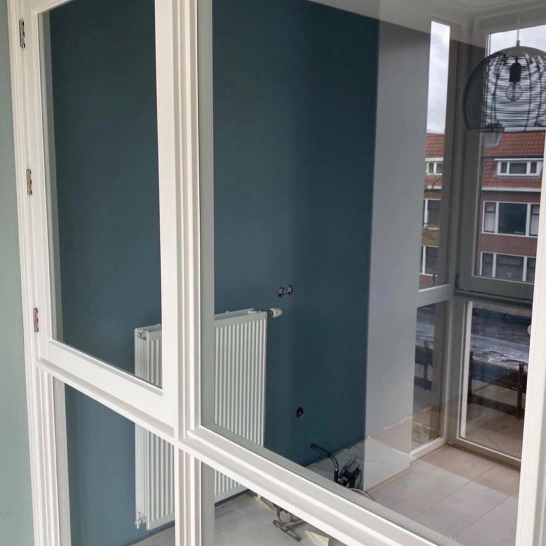 Een strak geschilderde ruimte met een diepblauwe accentmuur en friswitte kozijnen, vastgelegd door het glas van een binnendeur; de moderne afwerking, de witte radiator en het binnenvallende daglicht zorgen voor een frisse, eigentijdse uitstraling.