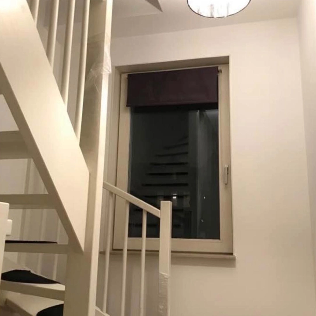 Een helder wit geschilderde trapopgang met strakke leuningen en een raam dat uitzicht biedt op een moderne zwevende trap aan de andere zijde, subtiel verlicht door een plafondlamp – een combinatie van functionaliteit en eigentijdse afwerking.