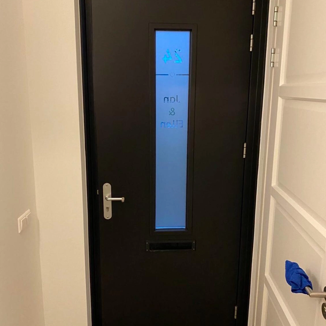 Deze zwarte voordeur met smalle, verticale glasstrook straalt kracht en elegantie uit. De matte afwerking en het strakke lijnenspel combineren mooi met het witte interieur, terwijl het geëtste glas zorgt voor privacy én een subtiele persoonlijke touch – hier zelfs met namen en pictogrammen. Een stijlvol welkom!