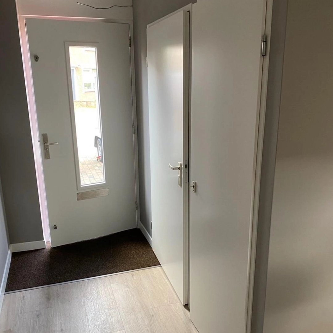 Fris geschilderde hal met witte binnendeuren, een witte voordeur met smal raam en brievenbus, lichtgrijze muren en een lichte houten vloer, die samen zorgen voor een moderne, strakke uitstraling.