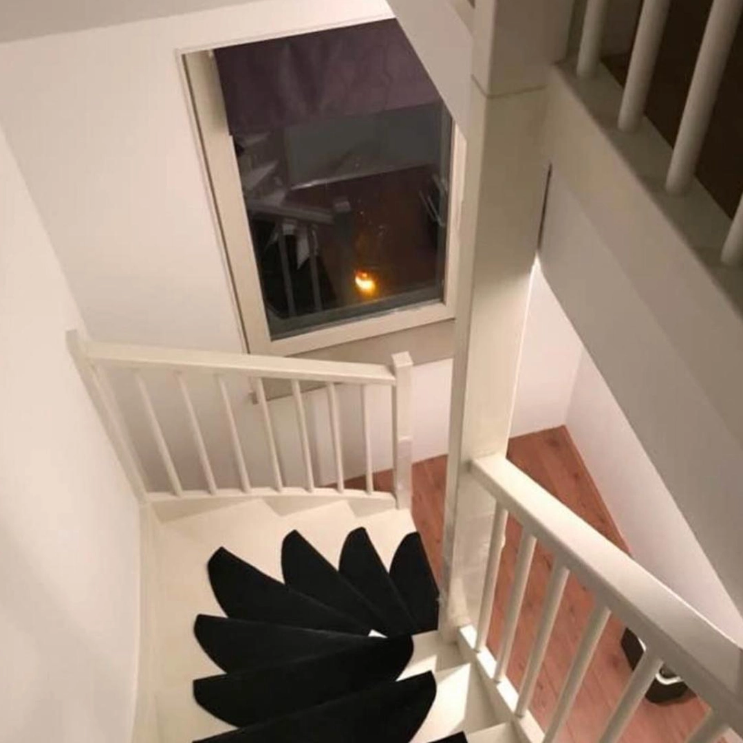 Trapgat met wit geschilderde trap en balustrade, voorzien van zwarte antislip-traptreden, witte wanden en een houten vloer op de benedenverdieping; door het raam is de trap weerspiegeld zichtbaar.