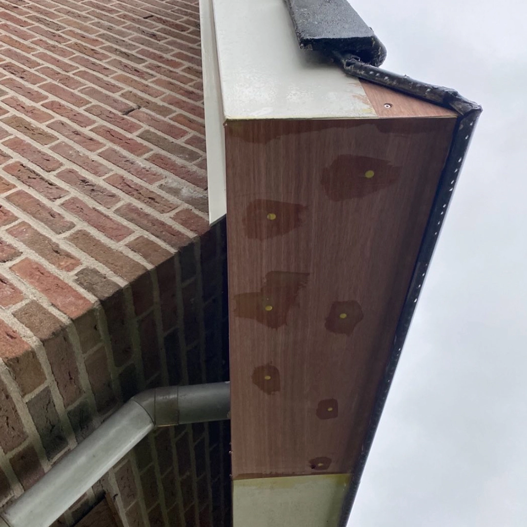 Herstel van een houten dakgootafwerking waarbij nieuwe multiplexplaat is aangebracht; schroefgaten zijn dichtgezet met houtvuller, en de overgang naar het witte boeiboord is netjes afgekit; links is een bakstenen gevel zichtbaar met een zinken regenpijp.