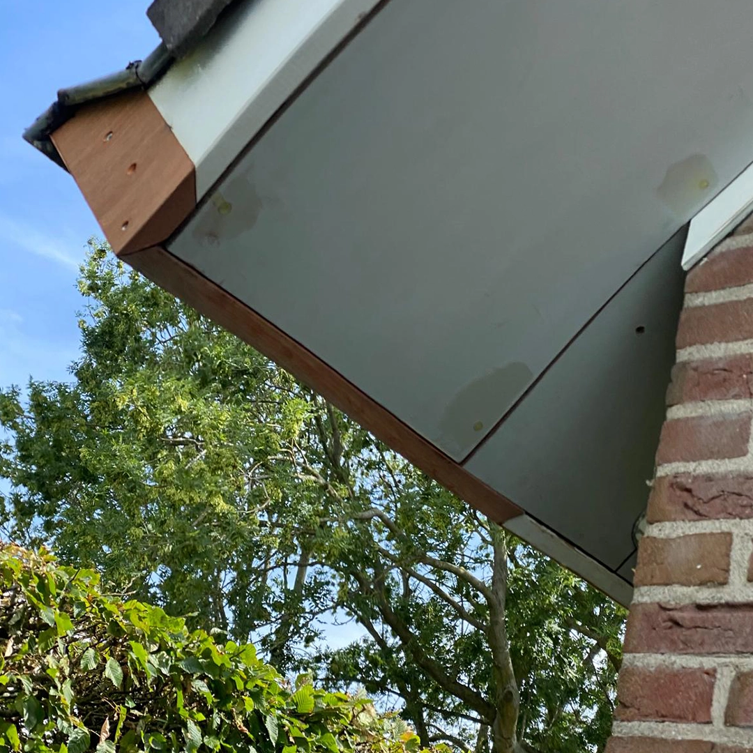 Herstel van dakgootbetimmering waarbij nieuwe houten delen zijn aangebracht en de naden en schroefgaten zijn geplamuurd, met op de achtergrond een blauwe lucht, groene bomen en een gemetselde gevel.