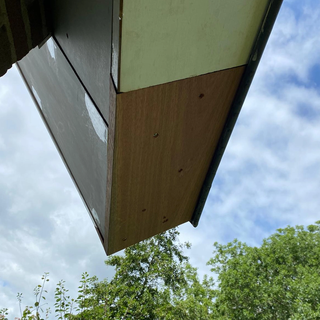 Herstelwerk aan een dakgootrand waarbij nieuwe multiplex platen zijn geplaatst en bevestigd met schroeven; het oude houtwerk aan de zijkant is deels gerepareerd en geplamuurd, met op de achtergrond een deels bewolkte hemel en groene bomen.