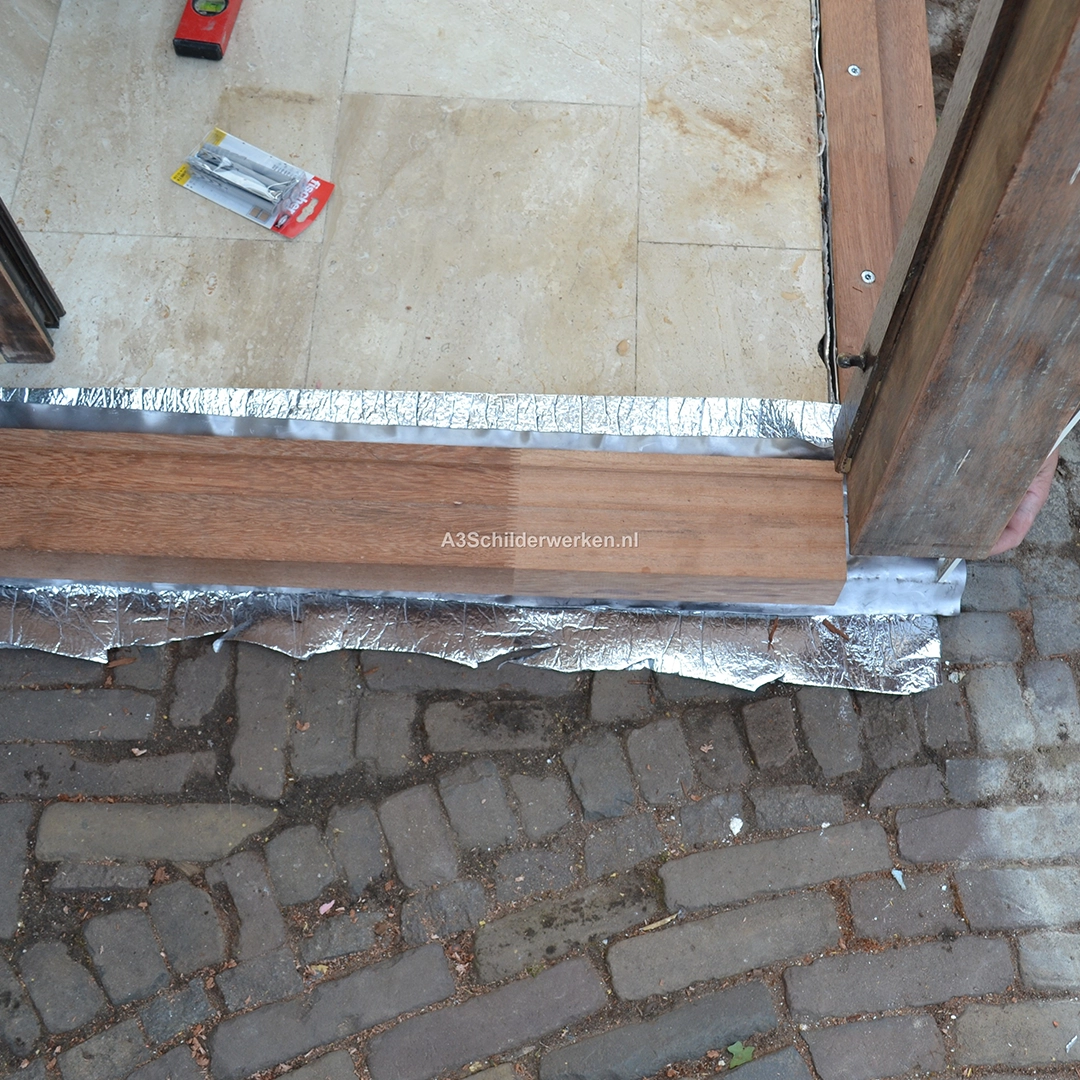 Herstel van een houten onderdorpel in een buitendeurkozijn, waarbij nieuw hout is geplaatst op een vochtwerende folie over een natuurstenen vloer; schroeven en gereedschap zichtbaar, duidend op lopende renovatiewerkzaamheden.