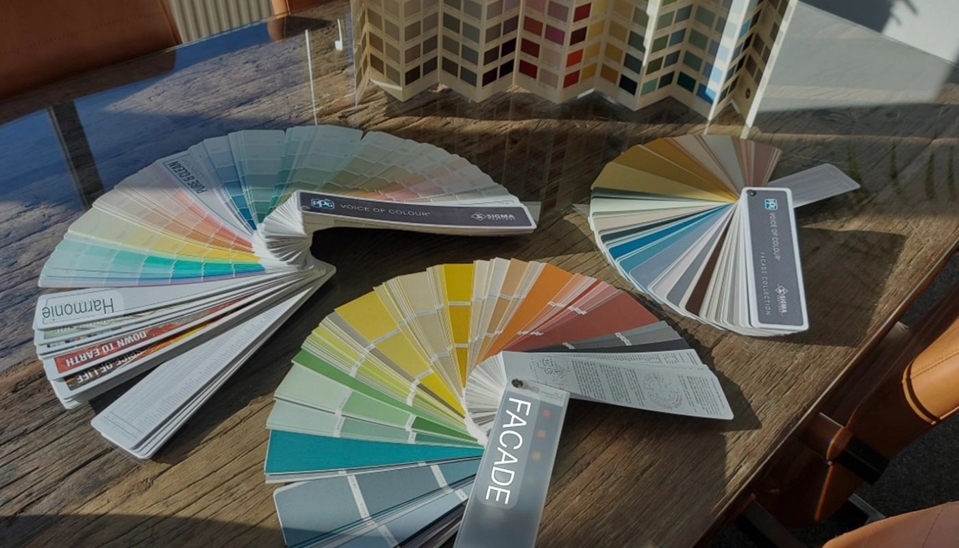 Op een houten tafel liggen verschillende kleurstalenwaaiers van Sigma Coatings opengevouwen, elk met een breed spectrum aan kleuren voor verf- en interieurkeuze; op de achtergrond staat een driedimensionaal kleurpalet en zijn leren stoelen en zonlicht zichtbaar.