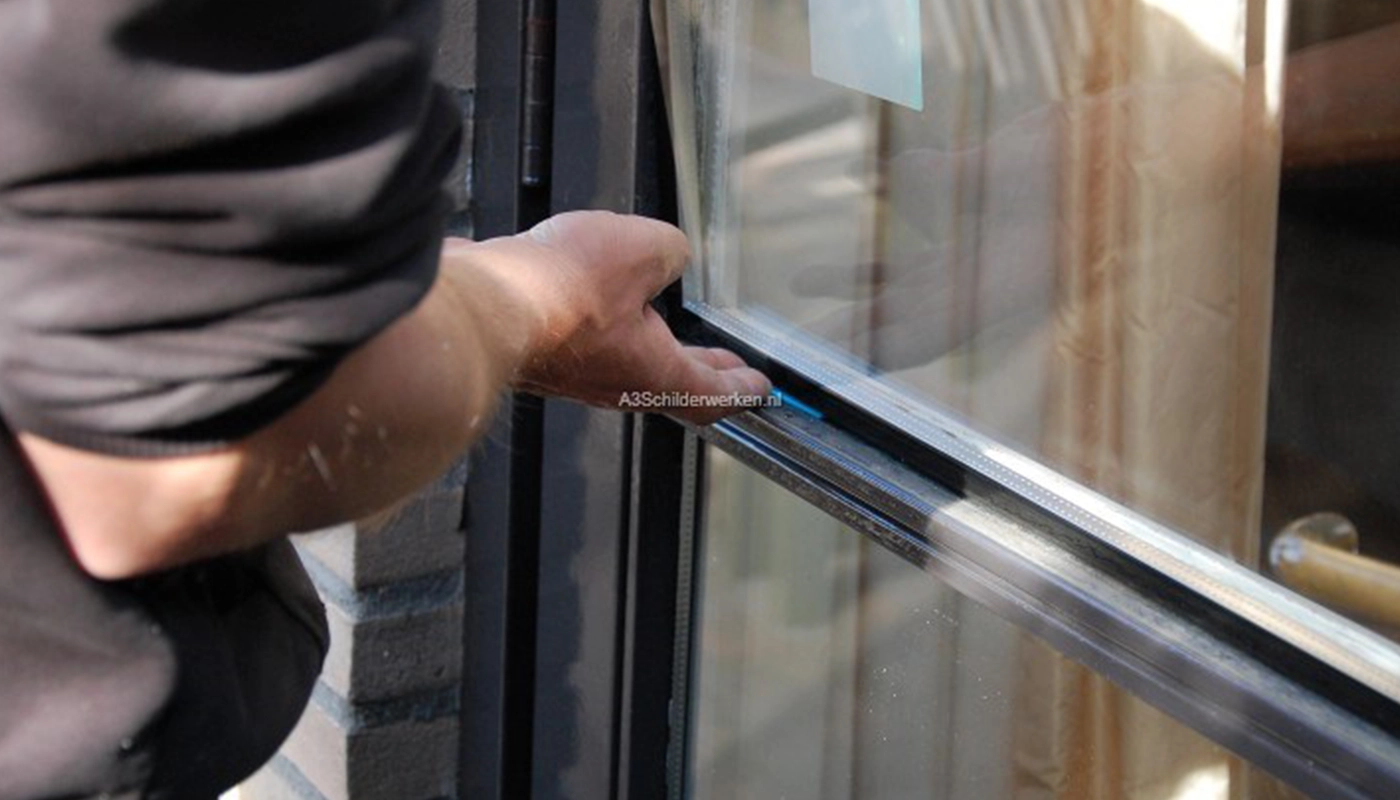 Een schilder of glaszetter plaatst zorgvuldig een ruit in een kozijn, waarbij zijn hand het glas op zijn plek houdt; op de achtergrond zijn een bakstenen gevel en de weerspiegeling van gordijnen zichtbaar.