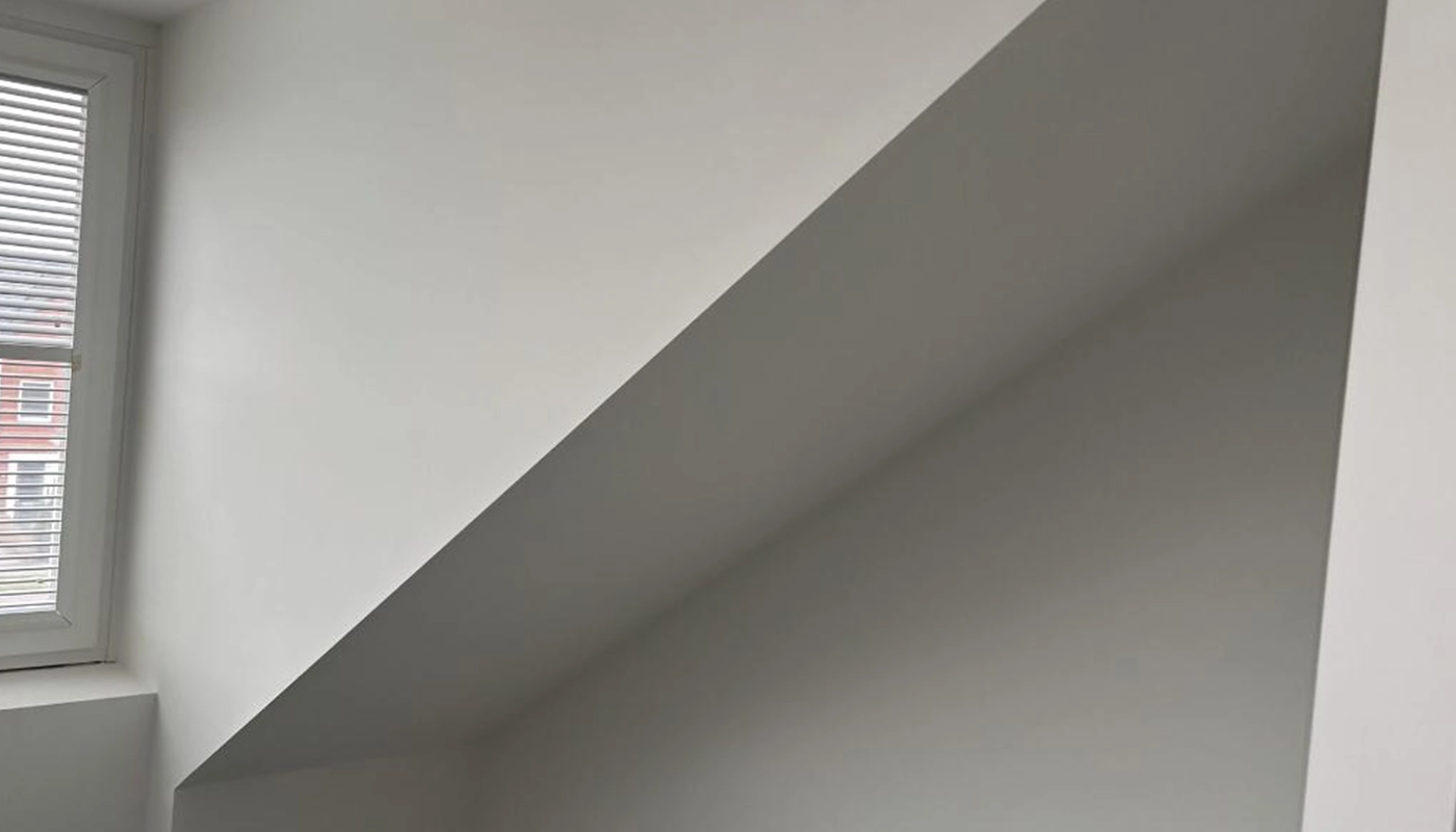 Een strak afgewerkte binnenmuur met renovliesbehang in een ruimte met een schuin plafond, naast een raam met witte jaloezieën waar daglicht naar binnen valt.