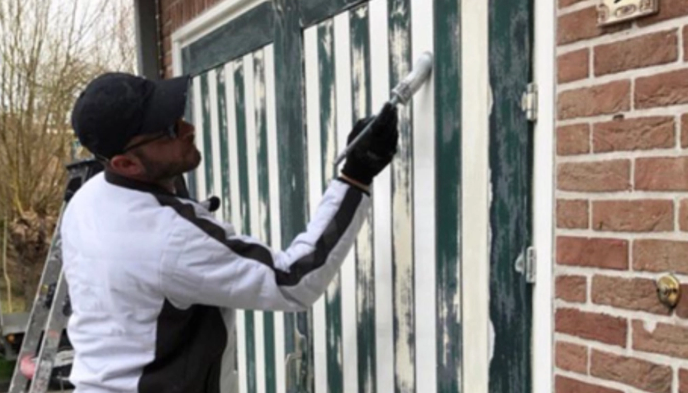 Een schilder in een wit met zwart schildersjack en pet werkt nauwkeurig aan het schilderen van een groen-wit gestreepte garagedeur; op de achtergrond is een stenen gevel en een ladder zichtbaar.