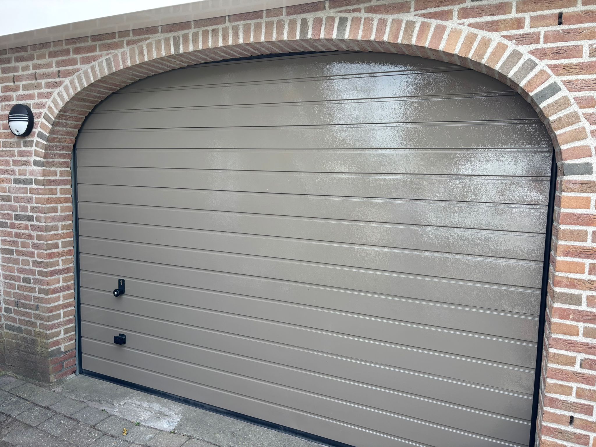 Een grijs bruine garage deur die net geschilderd is. Daaromheen een steenwerk in de vorm van een boog.