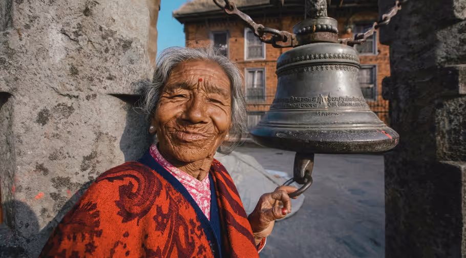 Femme âgée népalaise vêtue de rouge sonnant une cloche devant un temple.