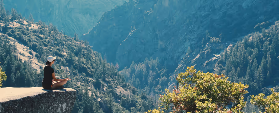 Femme méditant au bord d'un rocher face aux montagnes et aux arbres.